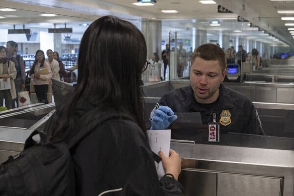 A U.S. border agent wearing a dark uniform sits behind a metal counter at an airport. He is wearing a light blue glove as he prepares to accept a document from a traveler with long, black hair who is facing him across the counter. A U.S. border agent wearing a dark uniform sits behind a metal counter at an airport. He is wearing a light blue glove as he prepares to accept a document from a traveler with long, black hair who is facing him across the counter.