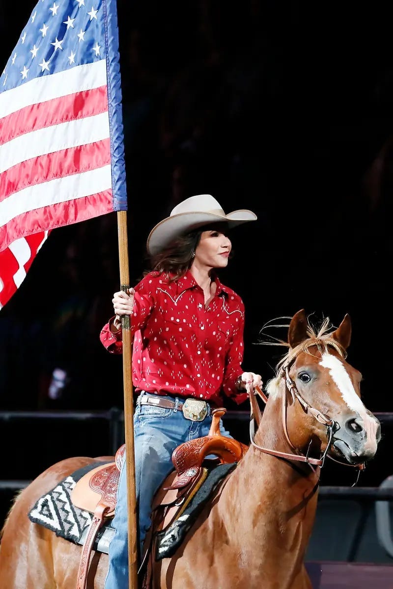 Kristi Noem, in a cowboy hat, Western shirt and jeans, holds an American flag while riding a horse. Kristi Noem, in a cowboy hat, Western shirt and jeans, holds an American flag while riding a horse.