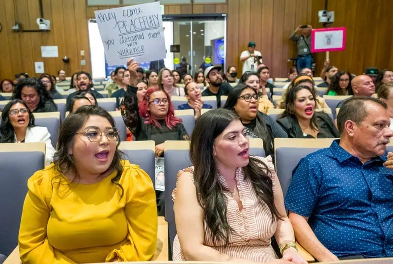 People sitting in a large room chanting. One person is holding up a sign that says “they came peacefully you detained them with violence.” People sitting in a large room chanting. One person is holding up a sign that says “they came peacefully you detained them with violence.”