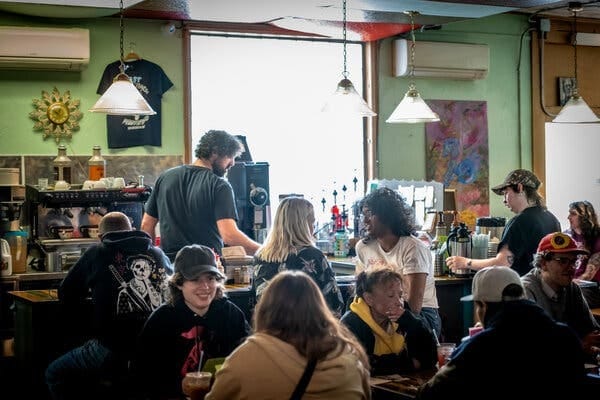 A cafe scene with people talking and interacting. Staff work behind a counter with a coffee machine, while customers sit at tables. A cafe scene with people talking and interacting. Staff work behind a counter with a coffee machine, while customers sit at tables.