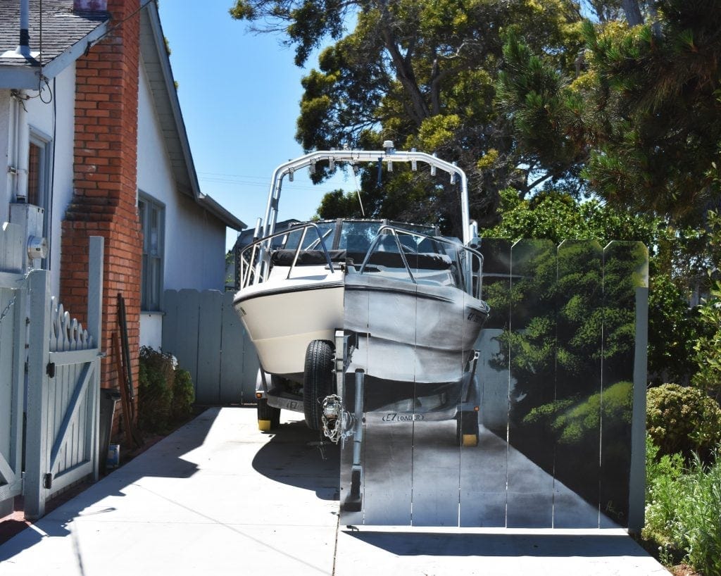 A photograph showing a boat in a driveway behind a fence with a boat painted on it A photograph showing a boat in a driveway behind a fence with a boat painted on it