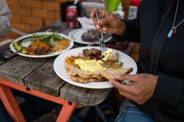 A person lifts yellow scrambled eggs with a fork. Hash browns, toast, and meat are on their plate, on a wooden table. A person lifts yellow scrambled eggs with a fork. Hash browns, toast, and meat are on their plate, on a wooden table.
