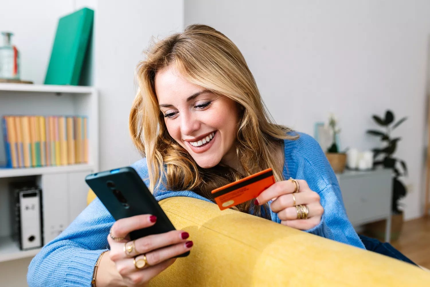 Similing young woman holding a phone and a credit card while sitting on a yellow couch. Similing young woman holding a phone and a credit card while sitting on a yellow couch.