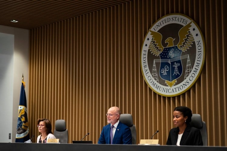 Three people, two women and a man, sit at a long desk in front of microphones. Behind them is the Federal Communications Commission logo. Three people, two women and a man, sit at a long desk in front of microphones. Behind them is the Federal Communications Commission logo.