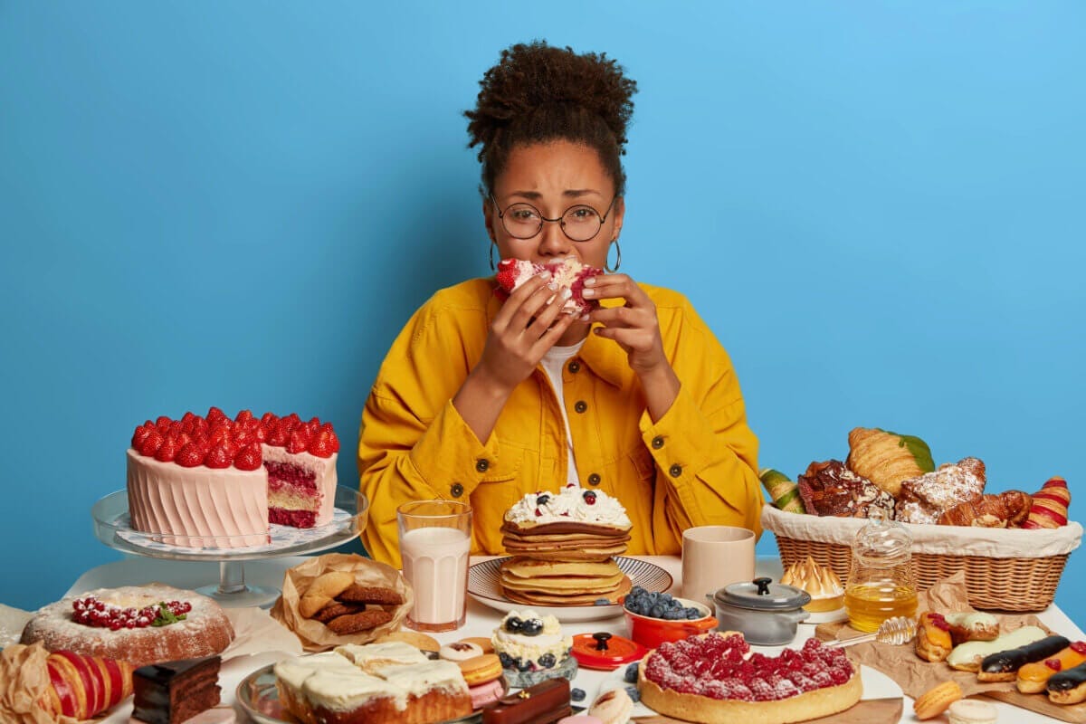 Gluttony and overeating concept. Upset crying ethnic woman eats piece of cake reluctantly, sits at table with many desserts, isolated over blue wall, feels hungry and greedy, wears yellow jacket Gluttony and overeating concept. Upset crying ethnic woman eats piece of cake reluctantly, sits at table with many desserts, isolated over blue wall, feels hungry and greedy, wears yellow jacket