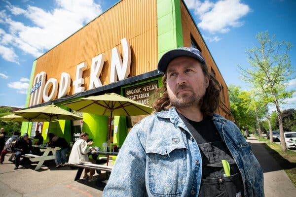 A person in a blue cap and denim jacket stands in front of a bright orange restaurant with lime green trim. Diners sit at picnic tables under green umbrellas in the midground. A person in a blue cap and denim jacket stands in front of a bright orange restaurant with lime green trim. Diners sit at picnic tables under green umbrellas in the midground.