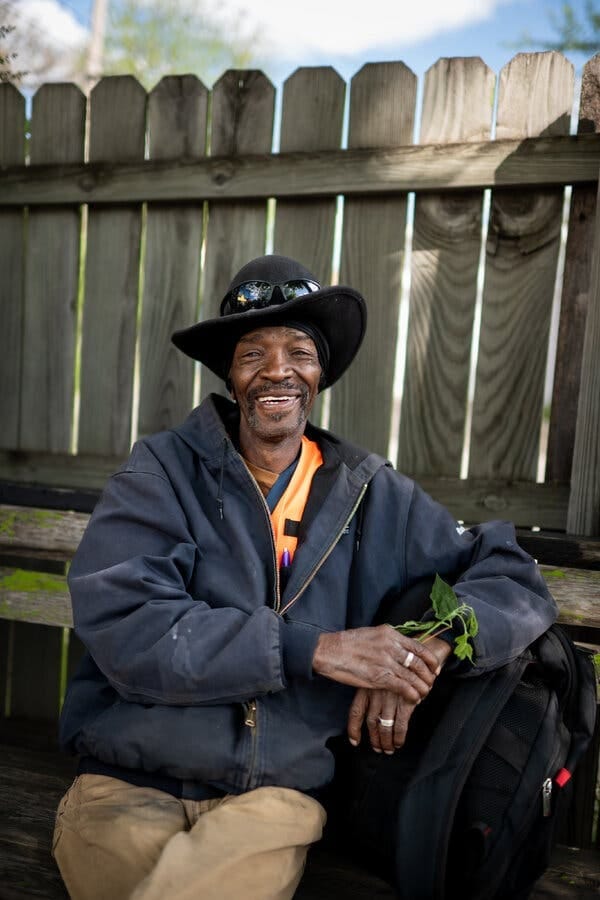 A man in a black hat, dark jacket, and orange shirt sits on a wooden bench. He is smiling and holding a small green plant. A wooden fence is in the background. A man in a black hat, dark jacket, and orange shirt sits on a wooden bench. He is smiling and holding a small green plant. A wooden fence is in the background.