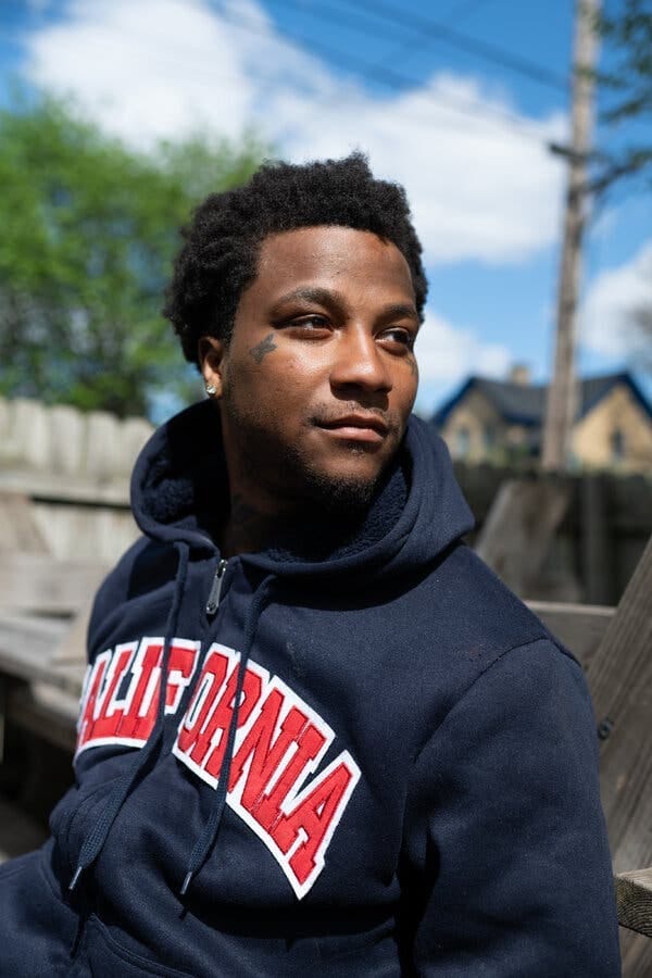 A young man with dark, curly hair and a tattoo near their eye wears a blue sweatshirt with red lettering. He is seated outdoors, with a wooden fence and blue sky behind him. A young man with dark, curly hair and a tattoo near their eye wears a blue sweatshirt with red lettering. He is seated outdoors, with a wooden fence and blue sky behind him.