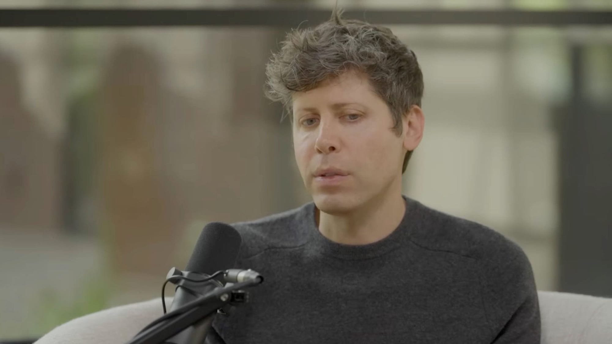 Altman with short curly brown hair wearing a dark gray sweater speaking into a black podcast microphone. Altman with short curly brown hair wearing a dark gray sweater speaking into a black podcast microphone.