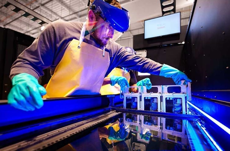a man wearing rubber gloves and a visor lowers a circuit board into a trough containing a liquid a man wearing rubber gloves and a visor lowers a circuit board into a trough containing a liquid