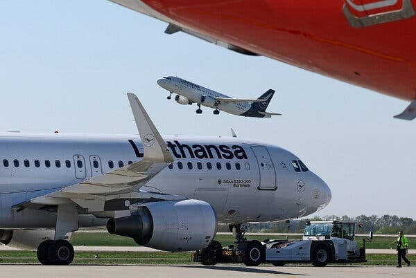 A red airplane wing is in the top foreground. A white Lufthansa plane is on the tarmac, and another Lufthansa plane takes off against a clear sky. A red airplane wing is in the top foreground. A white Lufthansa plane is on the tarmac, and another Lufthansa plane takes off against a clear sky.