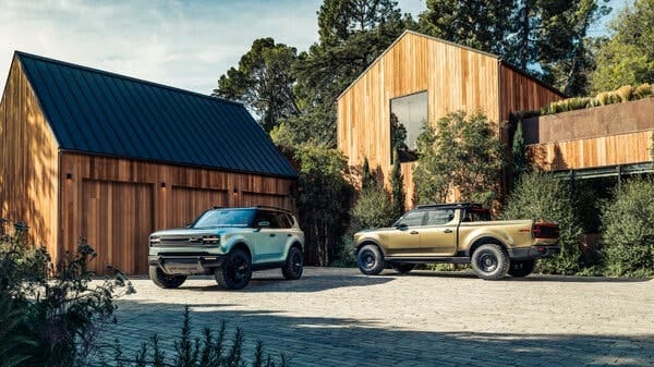A light-blue S.U.V. and a bronze-green pickup truck in front of a modern-looking house with elegant wood siding. A light-blue S.U.V. and a bronze-green pickup truck in front of a modern-looking house with elegant wood siding.