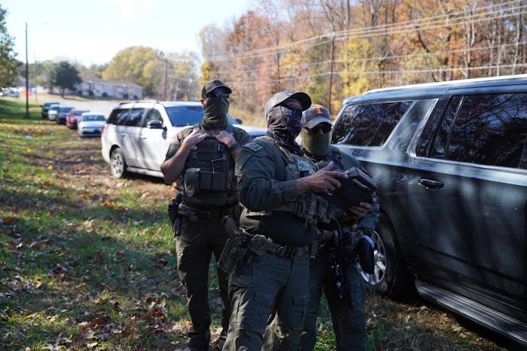 Masked officials stand next to a line of cars. Masked officials stand next to a line of cars.