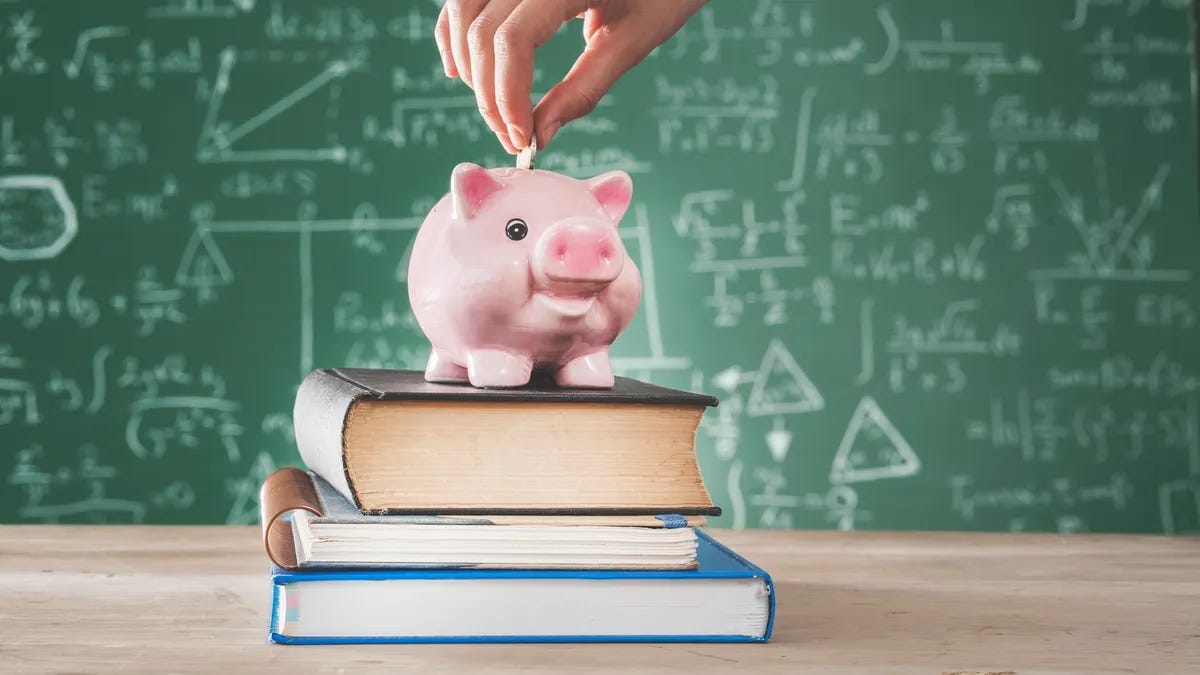 A stack of books is sitting on a table. A pink piggybank is on top of the stack of books. A hand is putting coins into the piggybank.