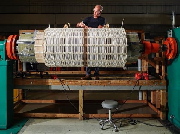 A person working at a large cylindrical piece of equipment that is wrapped in coils. A person working at a large cylindrical piece of equipment that is wrapped in coils.