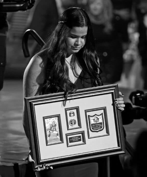 In a black-and-white image, Sydney Seau holds a frame with a patch for her father’s Hall of Fame induction and other mementos of his career.