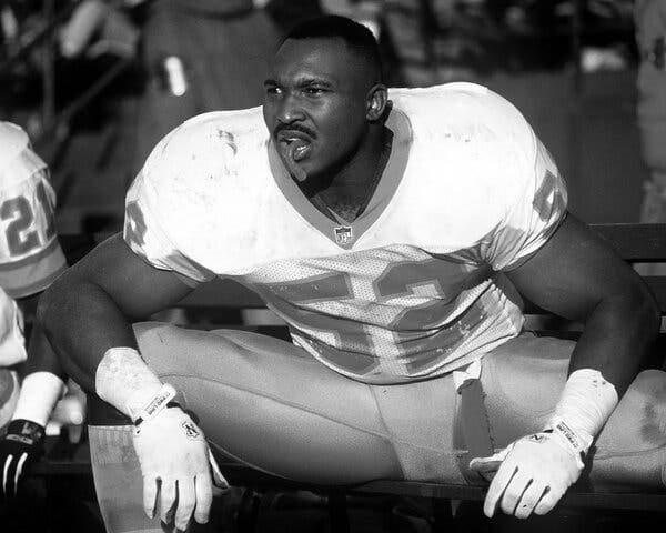 In a black-and-white photo, Keith McCants, sitting on a sideline bench, in a “52” football jersey and a mouth guard dangling out of his mouth. 