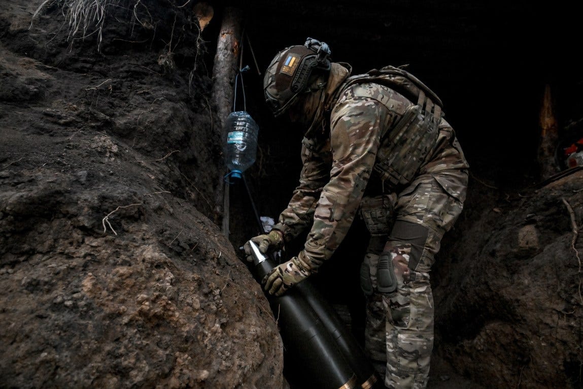 A soldier in a camouflage uniform handles 155 mm artillery shells, which are black with shiny, pointed tips.