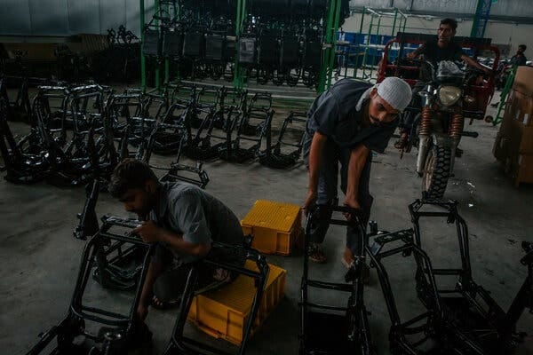 Two people work on black metal motorcycle frames lined up in rows on a concrete floor.