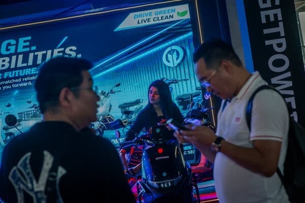 A woman sits on an electric motorcycle in front of a marketing poster that says “Drive Green Live Clean.”